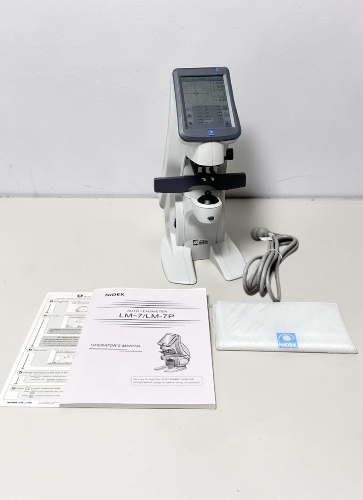 Nidek LM-7 auto lensmeter used — front view with operator's manual, dust cover and power cord